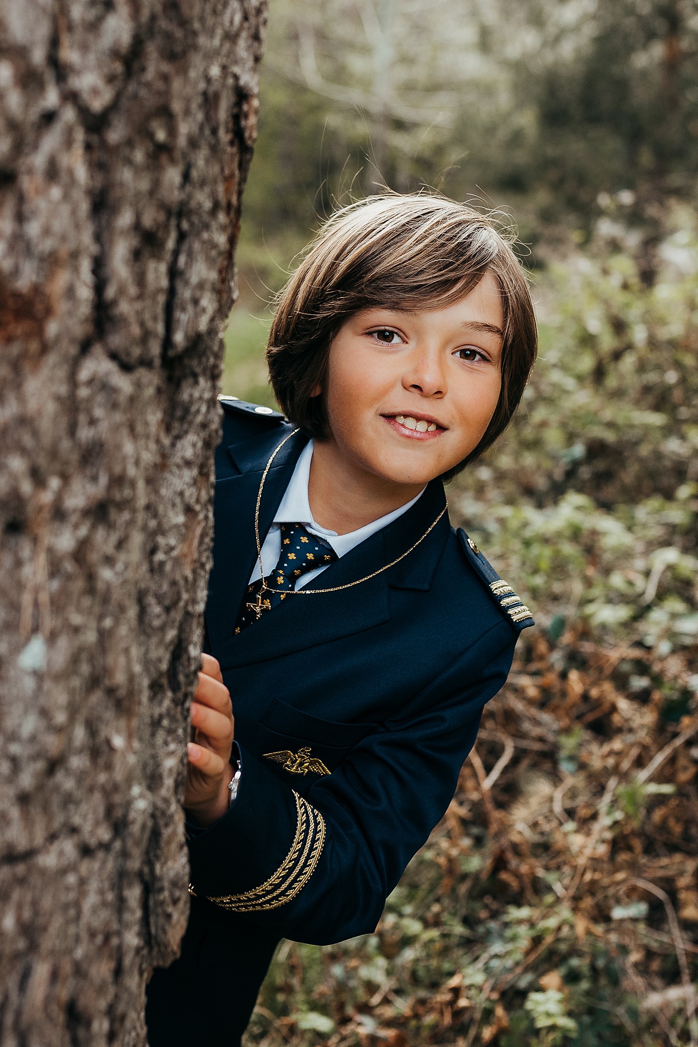 Niño con traje de comunion asomado detrás de un arbol al atardecer en una sesion fotografica natural al atardecer.